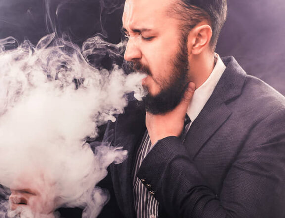 Ein Anzug tragender Mann mit Vollbartstößt mit Unbehagen auf dem Gesicht eine große Dampfwolke aus. Er fasst sich an den Hals und schaut auf die Dampfwolke.